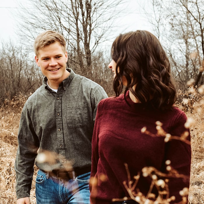 Engagement photographer - a couple smile at each other as they walk through a field