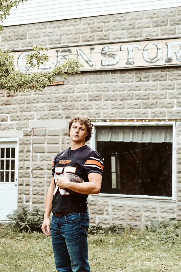 a boy poses in his football jersey, with a ball, for his senior portraits