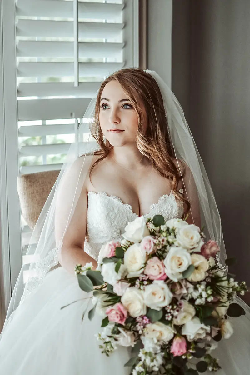 A bride holding her wedding bouquet sits on the edge of a bed, looking off to the distance