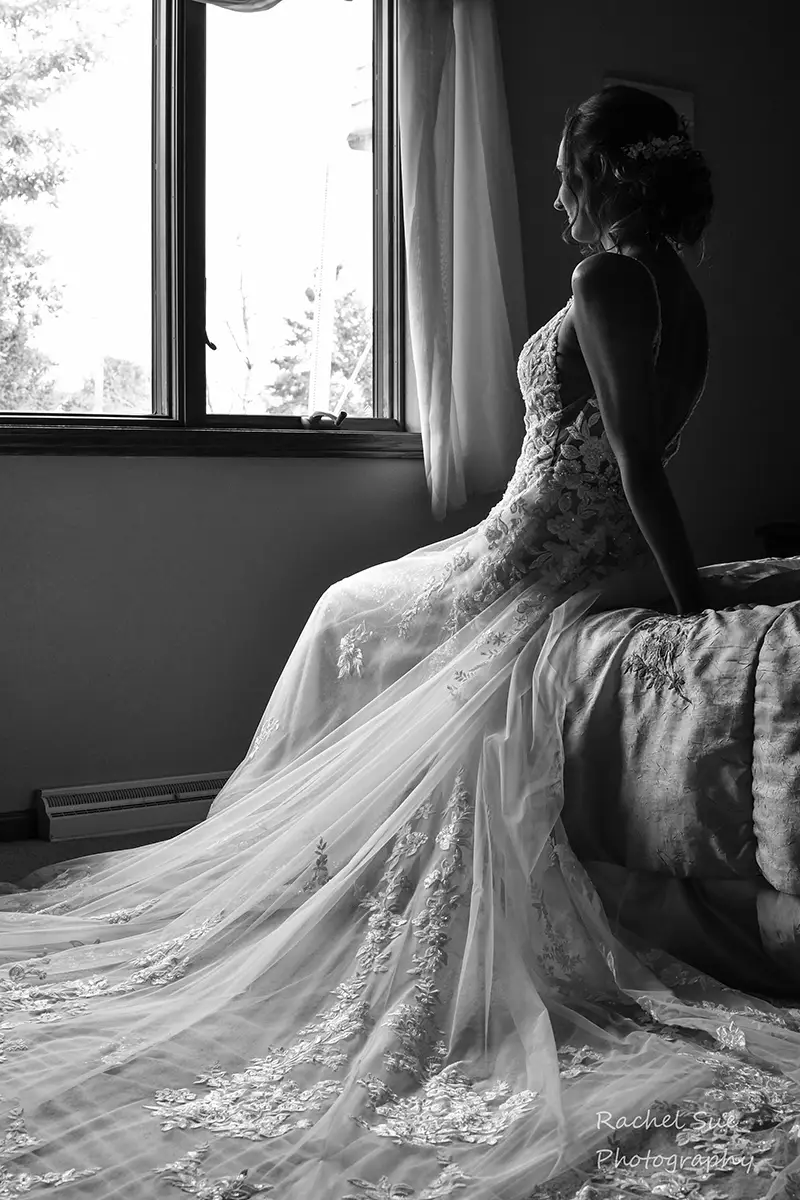 A black and white photograph of a bride sitting on the end of a bed, looking out of the window