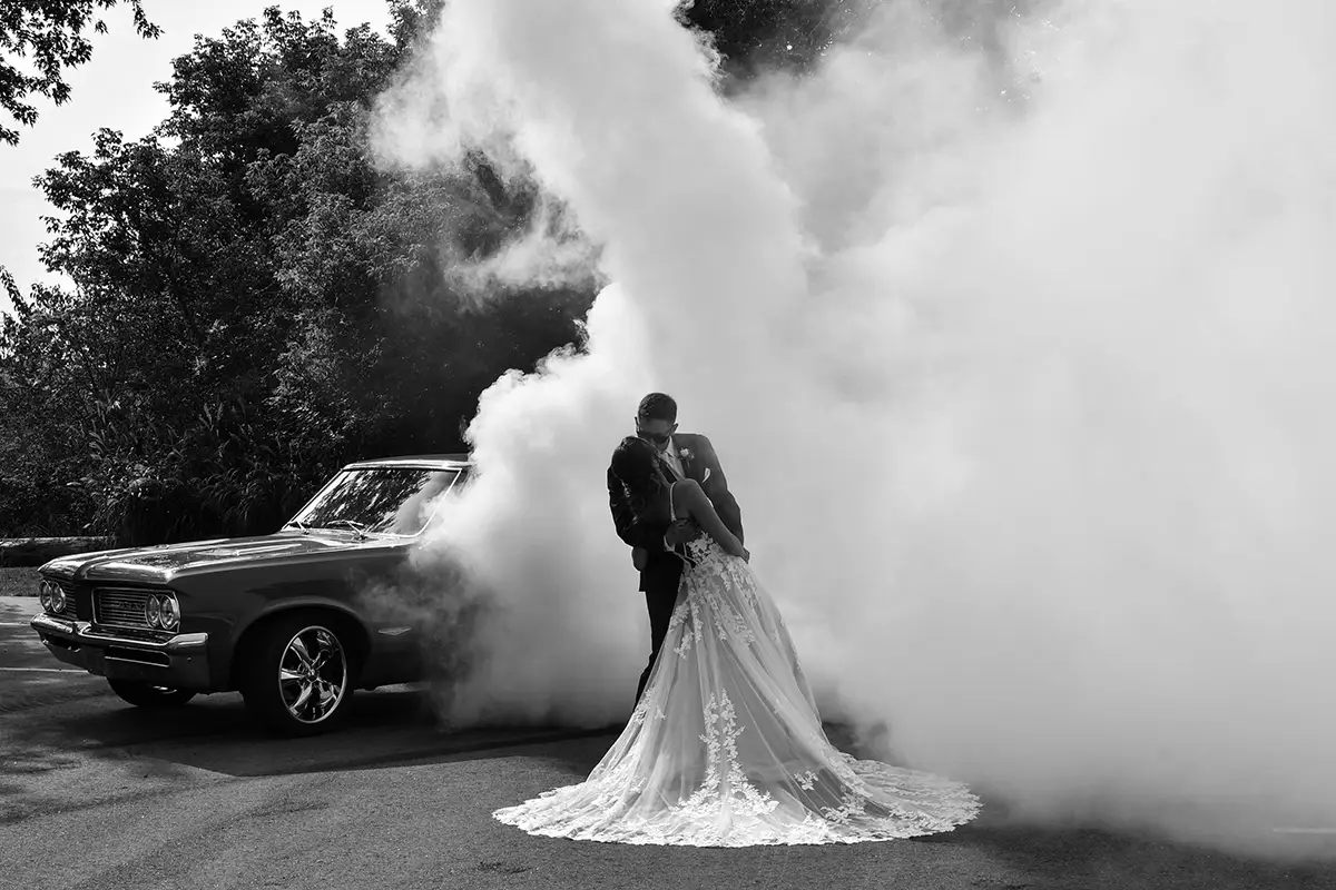 a bride and groom in front of their wedding car, surrounded by smoke for an atmospheric picture