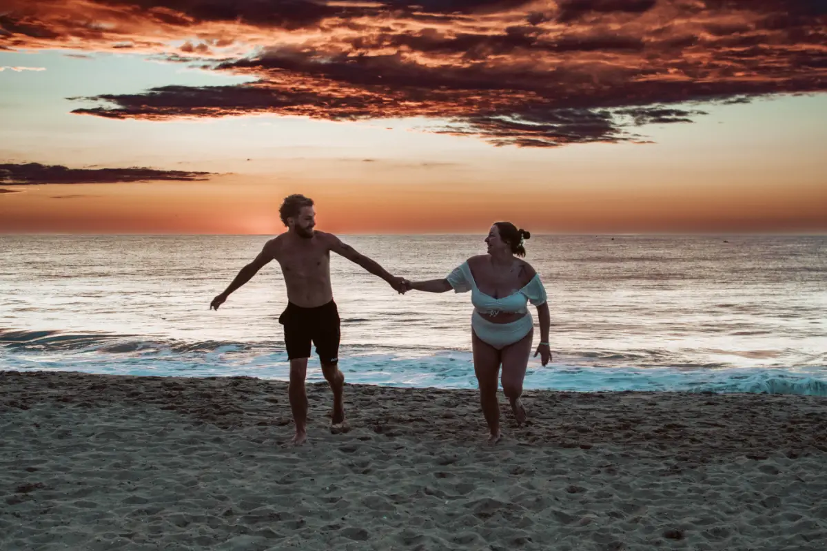 The newlyweds hold hands on the beach at sunrise. Behind them is a striking reddish black sky