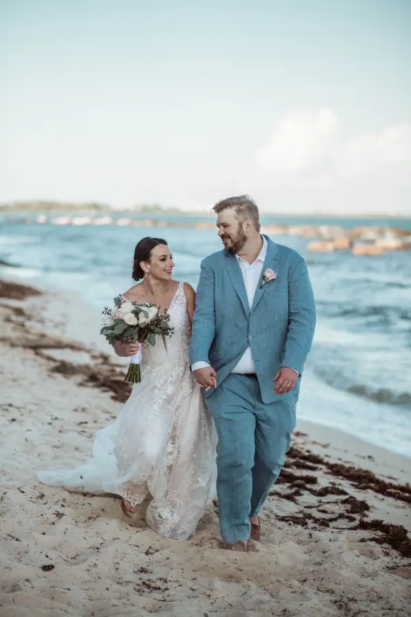 A newlywed couple walk hand-in-hand along the beach.