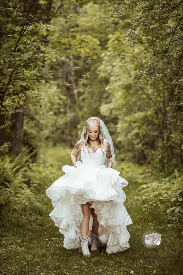 A bride, wearing a wedding gown, walks through the woods. She has hitched her skirt up to protect it from getting dirty, which reveals her white cowboy boots.